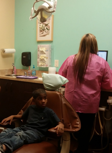 Young patient sits in dental chair while dentist in pink scrubs examines teeth in treatment room.